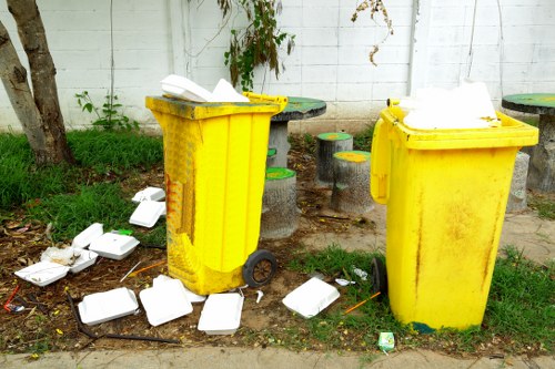 Two yellow wheelie bins with black wheels and closed lids are positioned on a sidewalk near a white-painted brick wall, with a small patch of grass and plants behind them. The bins are used for rubbish collection and are placed on a concrete surface. In front of and around the bins, there are numerous scattered white polystyrene takeaway containers, some open and others closed, with pieces spilling onto the ground. The containers are made of lightweight, textured foam and contrast with the bright yellow of the bins. The scene suggests a garden or local rubbish removal area, possibly in Norwood, with clutter indicating recent waste disposal. The environment is outdoor, illuminated with natural daylight, and features elements such as a small tree trunk on the left and some green foliage, emphasizing a typical street or garden edge where rubbish collection services by companies like Garden Clearance Norwood are often required for general waste clearing and rubbish removal tasks.