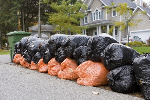 A row of large black and orange rubbish bags tightly filled and piled along the edge of a residential street, with some bags leaning slightly on each other and others resting directly on the pavement. The black bags occupy most of the foreground, showing a glossy, crumpled texture, while the orange bags are positioned beneath and partially visible, with a slightly softer appearance. Behind the bags, there is a suburban setting with a tree-lined pavement, a green wheelie bin, and a driveway leading to a modern, two-storey house with white and beige siding, multiple windows, and a parked car partially visible. The scene is illuminated by natural daylight, suggesting a clear or partly cloudy day, indicative of typical rubbish collection or garden clearance activity in Norwood, with the focus primarily on the accumulated waste for removal by Garden Clearance Norwood services.