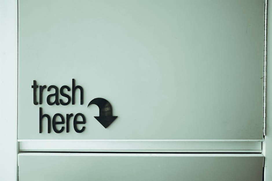 A narrow urban alleyway cluttered with a large gray and black recyclable waste container in the foreground, which appears to be made of fabric or composite material, with visible zippers and handles. 