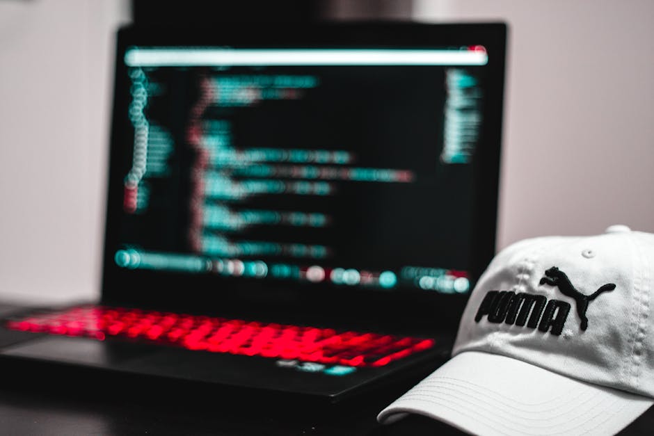 A person typing on a laptop computer with a dark-colored keyboard, displaying lines of source code on its screen. The screen shows a coding interface with various code blocks, highlighted syntax in gr